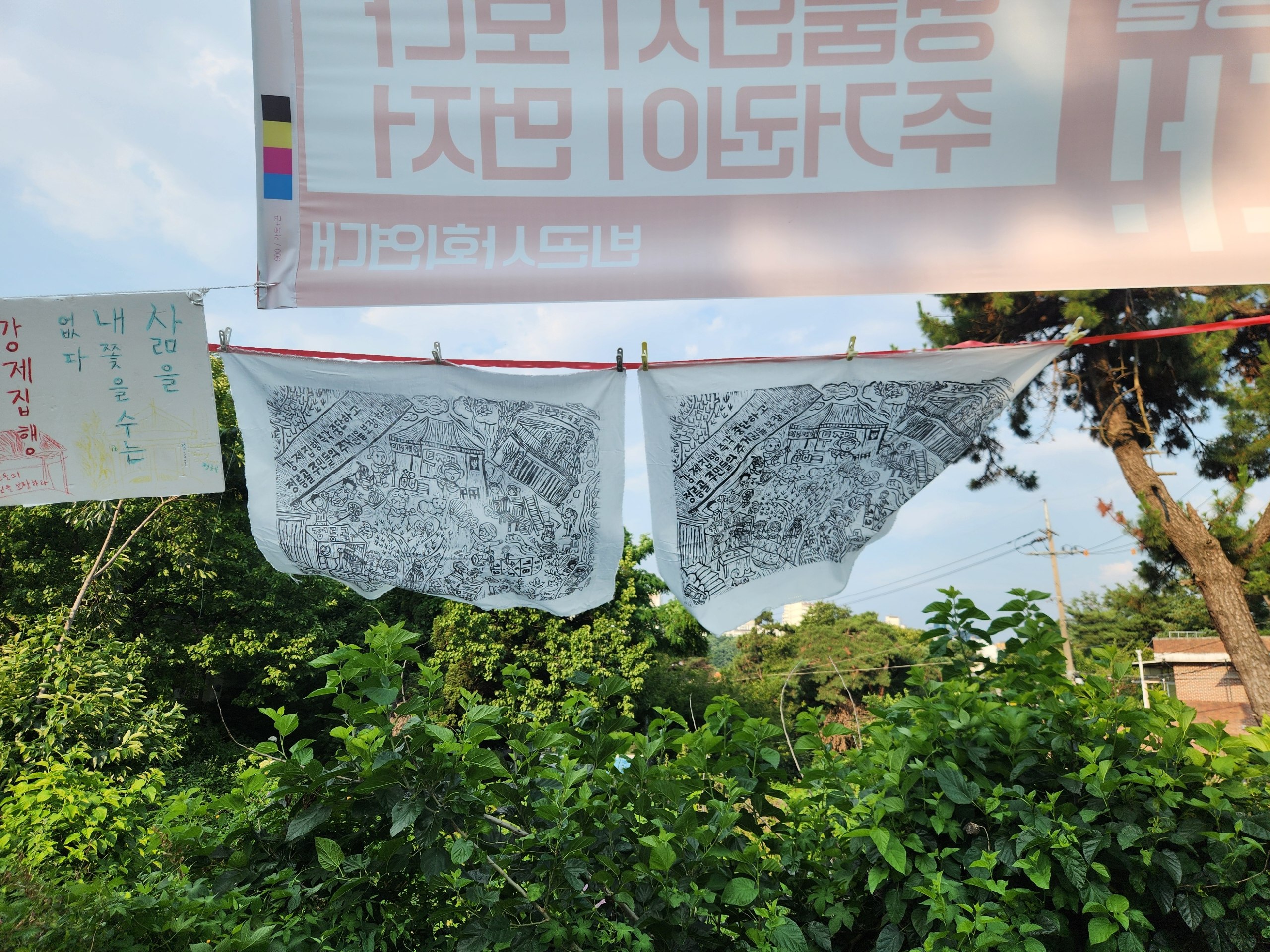 현수막 아래에 설치된 노끈에 판화와 작은 피켓이 매달려 바람에 날리는 모습을 촬영한 사진이다. 현수막은 뒷면에서 보이는 모습이고 '주거권이 먼저' 등의 문구가 적혀있다. 피켓에는 '삶을 내쫓을 수는 없다' 등의 문구와 그림이 그려져있다. 너머로는 수풀이 우거져있고 멀리 집과 전봇대가 보인다. 