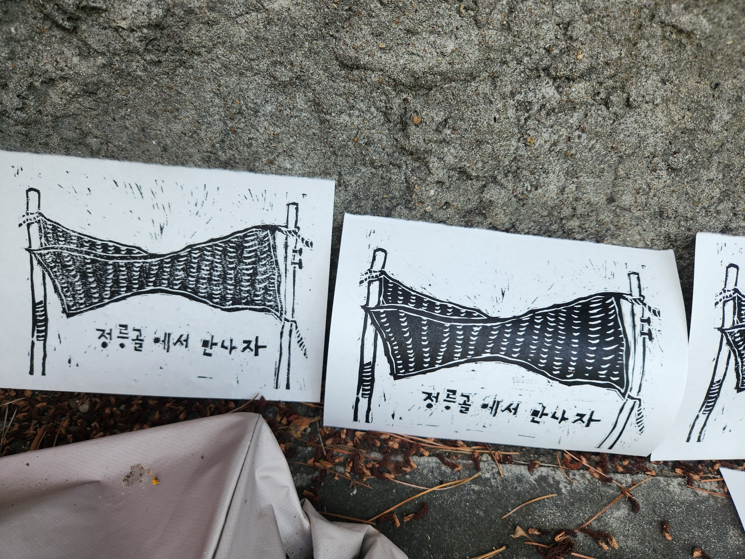 판화를 담벼락에 세워놓고 촬영한 사진이다. 두 기둥 사이에 묶인 배너의 모습, '정릉골에서 만나자'라는 문구가 판화에 표현되어 있다.