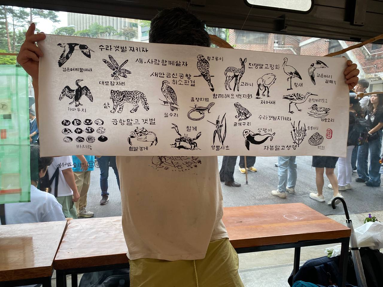 큰 유리창이 있는 실내 공간에서 한 사람이 좌우로 넓은 흰색 천에 찍힌 실크스크린 판화를 들고 서 있는 모습을 촬영한 사진이다. 판화에는 '수라갯벌 지키자. 새, 사람 함께살자. 새만금신공항 취소판결. 함께해요 백지화. 수라갯벌 지켜라. 공항말고 갯벌'등의 문구와 갯벌에 사는 여러 동식물들의 모습과 이름이 검은색으로 찍혀있다. 뒤에 유리창 너머로 바깥에는 여러 사람들이 서 있다. 