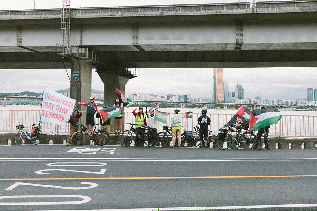 한강 공원 자전거 도로 옆에서 10여명의 사람들이 각각 자전거를 세워두고 팔레스타인 국기와 '이스라엘은 가자 학살 중단하라'고 적힌 깃발 등 다양한 연대 메시지를 담은 물품을 자전거에 매달고 정면을 바라보고 있는 모습을 촬영한 사진이다. 뒤로는 한강다리 진입로 구조물, 한강, 강 건너편의 풍경이 보이고 하늘은 구름이 많아 흐리다.