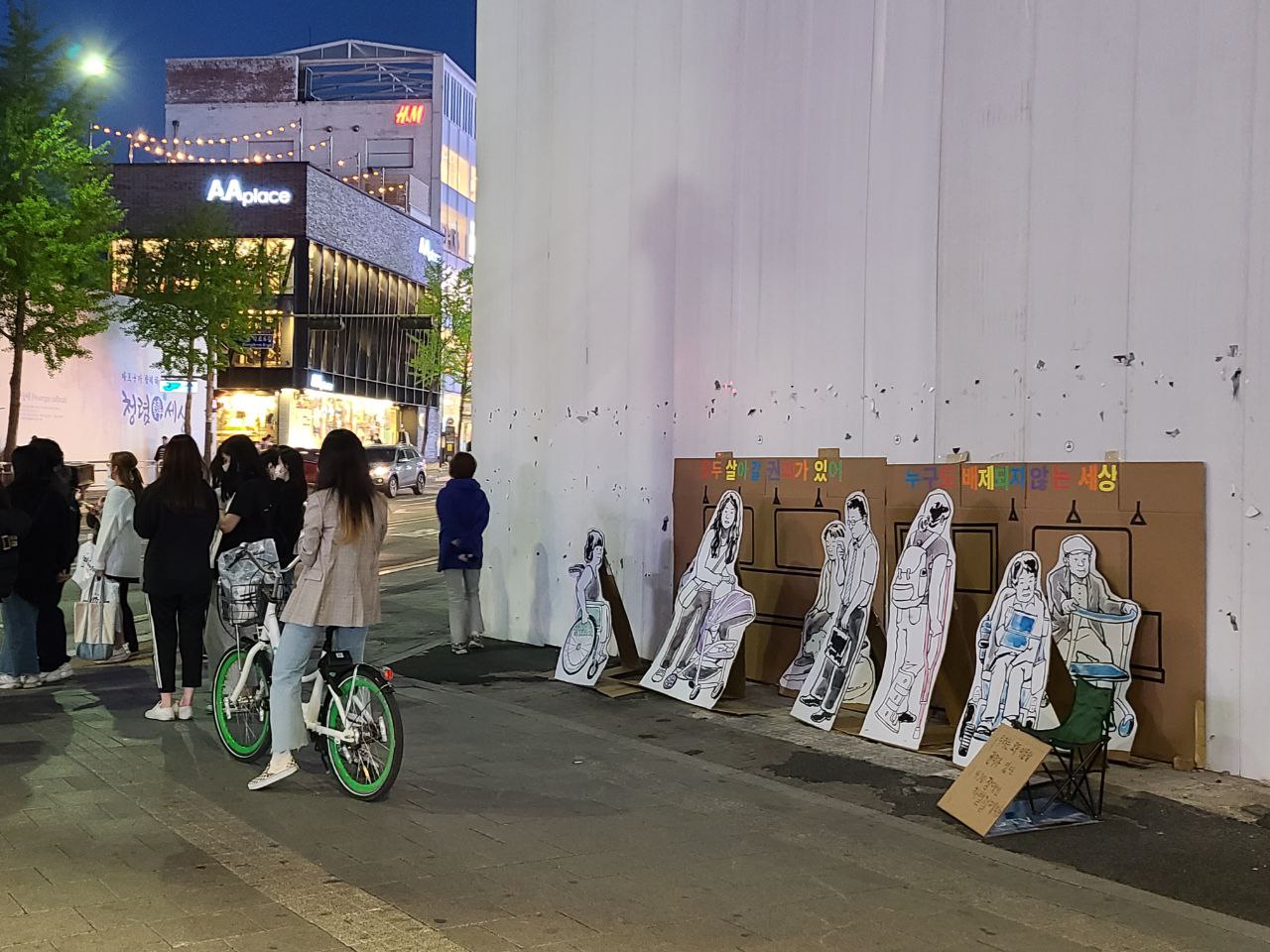 어두운 밤하늘과 가로등, 가로수. 불켜진 상가 건물이 왼쪽 너머로 보인다. 보행자 도로 위로 왼편에는 서 있는 사람들과 자전거를 탄 사람들이 있고, 오른편에는 높은 공사장 펜스가 있다. 공사장 펜스 앞에는 지하철을 표현한 박스 종이가 좌우로 넓게 세워져있다. 이 종이 위쪽에는 '모두 살아갈 권리가 있어. 누구도 배제되지 않는 세상'이라고 여러 색깔로 문구가 적혀있다. 박스 종이 앞에는 휠체어를 탄 사람, 아이를 데리고 있는 사람, 유아차를 모는 사람, 안내견을 동반한 사람, 휴대폰을 보며 서 있는 사람, 목발을 짚은 사람, 전동 휠체어를 탄 사람, 보행보조기를 쓰는 사람등의 모습이 그려진 종이가 세워져있다.