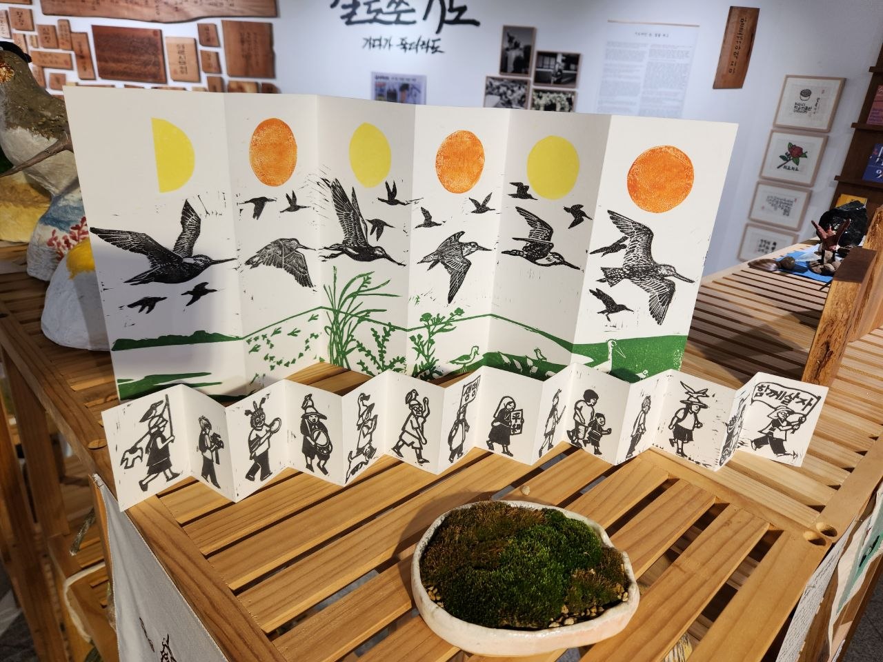 전시장 내부 선반을 가까이에서 촬영한 사진이다. 나무 선반 위에는 지그재그 형태로 접혀 세워진 종이에 찍힌 다색 판화가 가까이에 놓여있다. 뒤로는 여러 조형물이 보인다. 