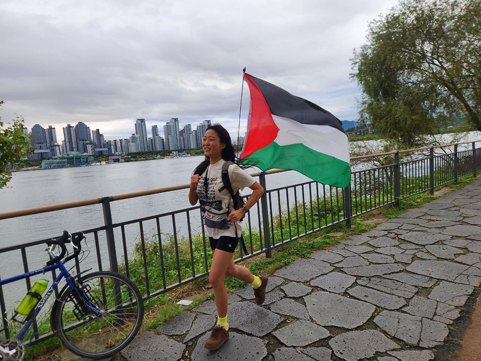 한강 공원 보행로 난간에 자전거를 세워둔 한 사람이 가방에 팔레스타인 국기를 부착하고 달리는 모습을 촬영한 사진이다. 강가에는 풀숲이 우거져 있고 강 건너편에는 높은 빌딩이 많이 보인다. 