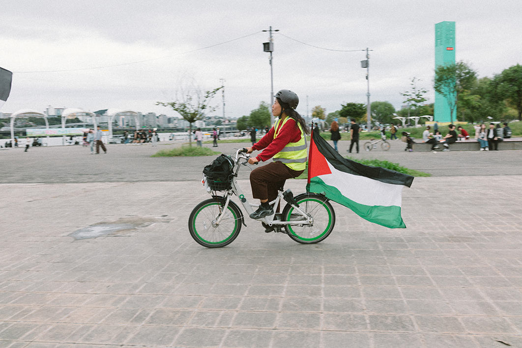  팔레스타인 국기를 매단 따릉이 자전거를 형광조끼와 헬멧을 착용한 사람이 타고 있는 모습을 촬영한 사진이다. 한강 공원 넓은 공터에서 자전거를 타고 있고 멀리 앉거나 걸어다니는 많은 사람들의 모습, 빌딩 등이 보인다. 