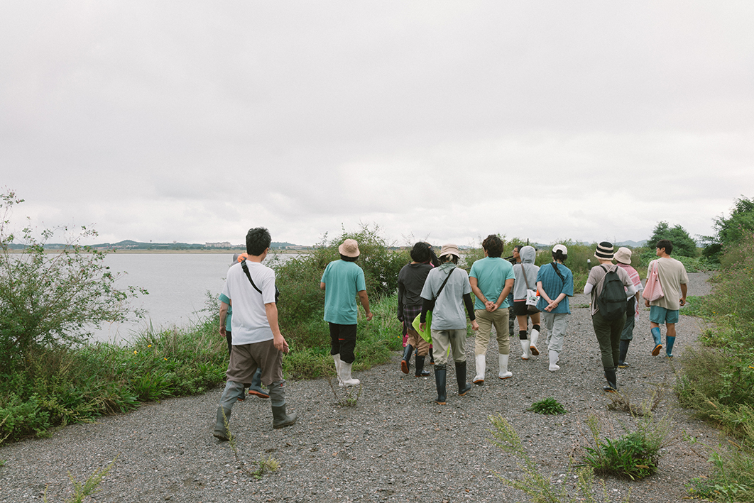 작은 자갈이 깔린 길을 약 13명의 사람들이 걸어가고 있는 모습을 촬영한 사진이다. 길 양 옆으로는 관목과 수풀이 우거져있고 왼쪽편으로는 바다와 멀리 육지의 모습이 보인다. 사람들은 장화를 신고 있으며 시선은 왼쪽 바다를 향해 있다. 하늘에는 구름이 많다.