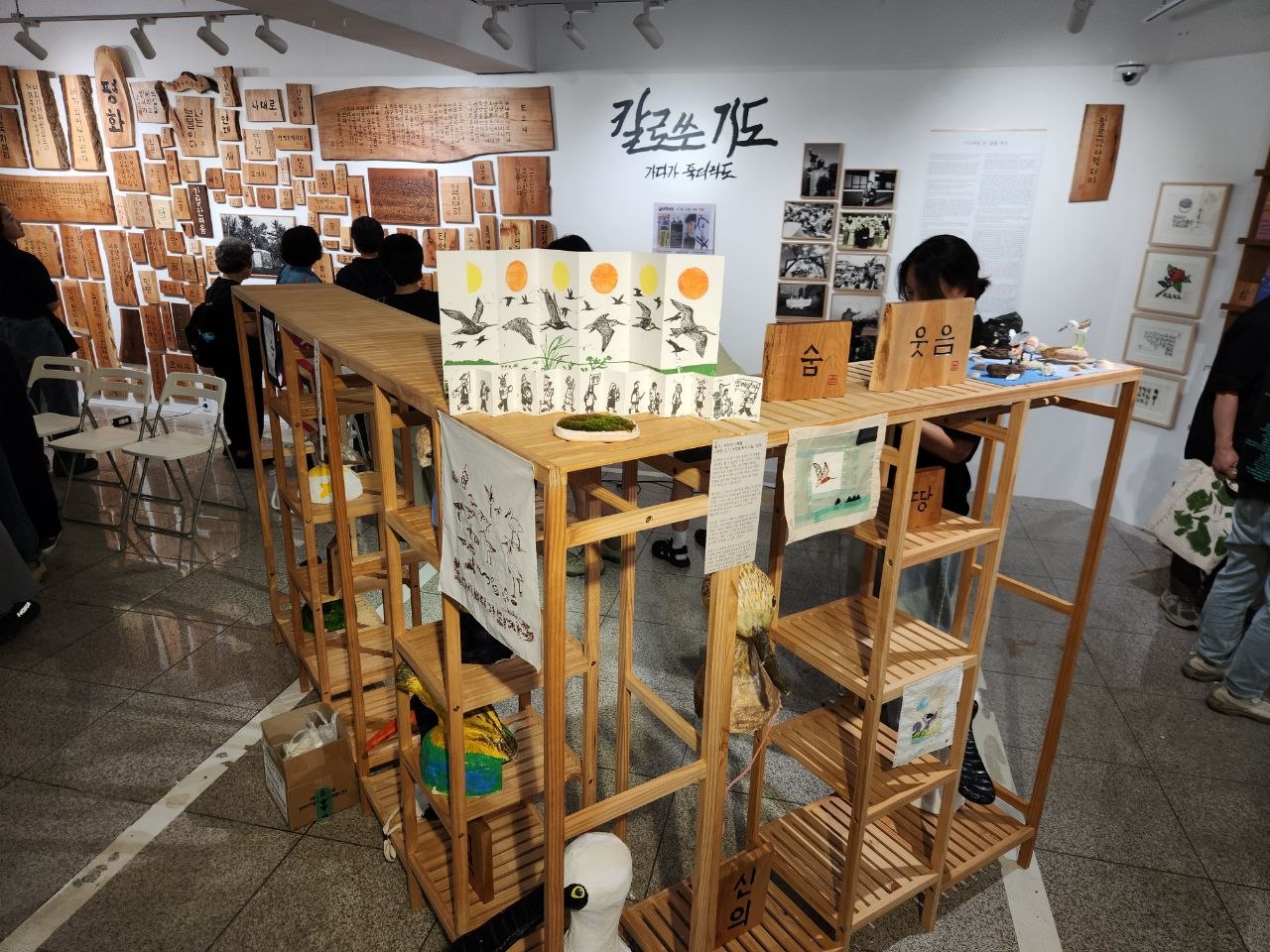 전시장 전경을 촬영한 사진이다. 가운데에는 기역 자 형태의 5단 나무 선반이 있고 각 칸마다 서각과 새모양 조형물 등이 배치되어 있다. 뒤쪽 벽면에는 나무 서각, 사진, 판화 등이 배치되어 있고 전시물을 보는 사람들이 약 9명이 서있다. 