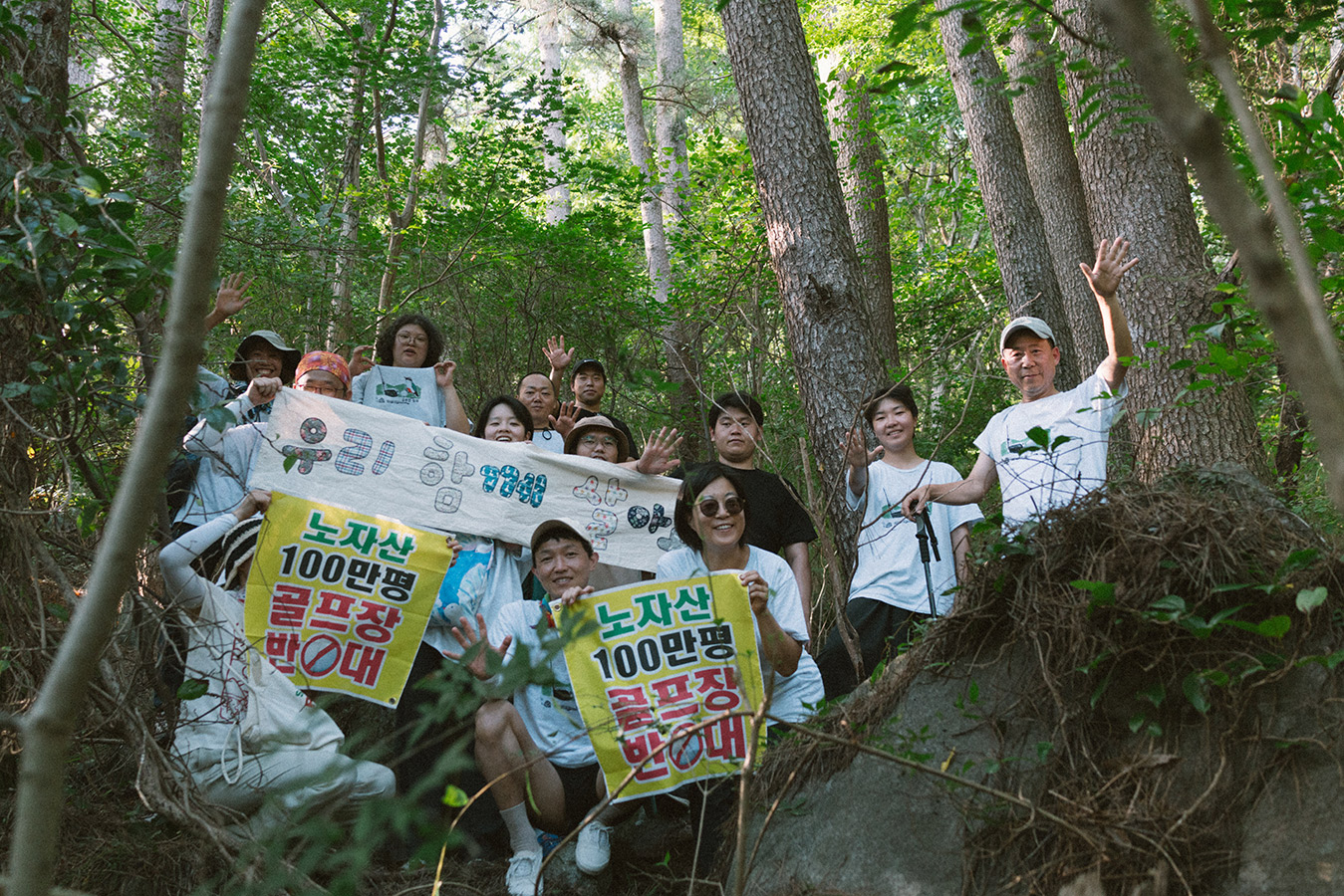 숲이 우거진 사이에 13명의 사람들이'노자산 100만평 골프장 반대, 우리 함께 살아요' 등의 문구가 적힌 다양한 현수막을 들고 서거나 앉아있다.