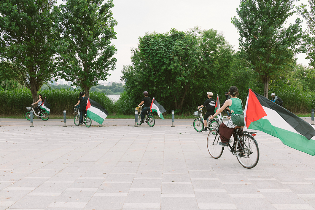 다섯 명의 사람들이 공터에서 한강 자전거길 방향으로 자전거를 타고 진입하고 있는 모습을 촬영한 사진이다. 자전거마다 팔레스타인 깃발이 부착되어 있다. 몇몇 사람들은 쿠피예를 두르고 있다. 뒤쪽으로 자전거를 타고 지나가는 사람이 한 명 있고, 강변으로는 수풀과 나무가 우거져있다. 