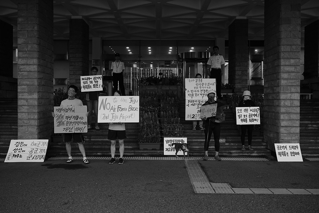 시설 입구 계단에 여섯명의 사람이 피켓을 들고 한 마리의 강아지와 함께 서 있는 장면을 찍은 흑백사진이다. 피켓에는 'No to the Jeju air force base (2nd Jeju airport)', '도민 위한 제2공항? 알고보니 공군기지! 원희룡 퇴진해!', '개발사업자가 셀프 작성 제출하는 셀프 환경영향평가제도 개선하라!' 등의 문구가 적혀있다. 뒤쪽 계단참에는 시설 직원으로 보이는 두 사람이 뒷짐을 지고 서 있다.