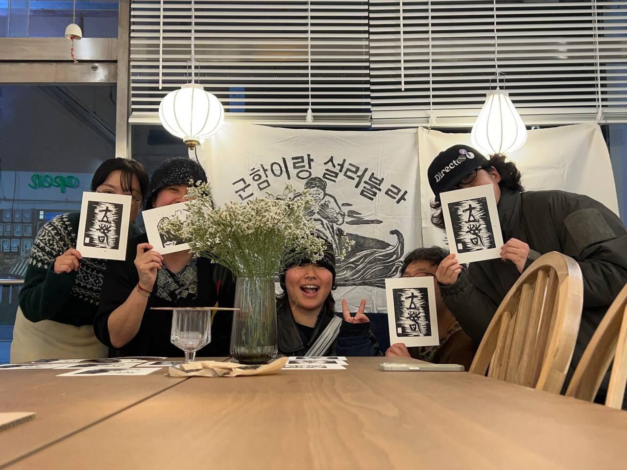 5명의 사람들이 실내의 테이블 옆에 앉거나 서서 판화를 들고 있는 모습을 촬영한 사진이다. 