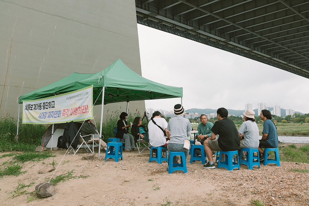 강가 다리 아래에 초록색 지붕의 천막이 설치되어 있고 주변으로 10여명의 사람들이 간이 의자를 이용해 둘러앉아 이야기를 나누는 듯한 모습을 촬영한 사진이다. 천막에는 '세종보 재가동 중단하고 4대강 재자연화 즉각 이행하라'라고 적혀있다. 천막 아래에는 낮은 텐트가 설치되어 있다. 천막이 있는 강변에는 고운 흙과 자갈이 있고 드문드문 풀이 자라있다. 뒤쪽으로 흐르는 강과 강 너머의 풍경이 보인다. 