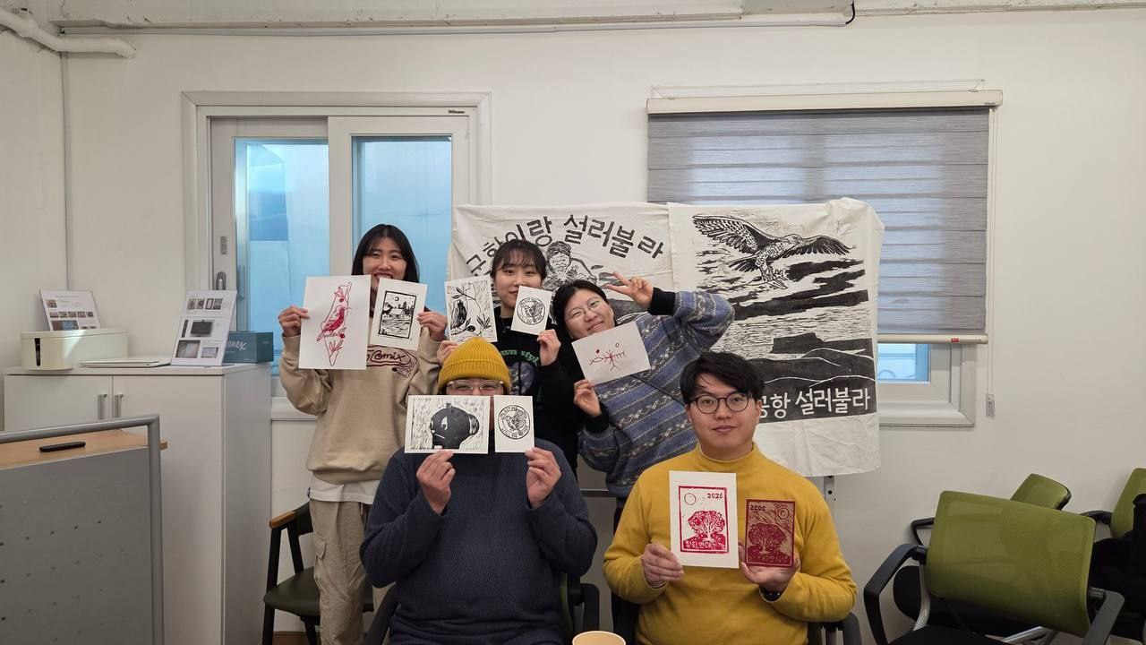 실내에서 5명의 사람들이 안거나 서서 판화를 들고 촬영한 사진이다. 뒤쪽 벽에는 작은 창 2개와 그 앞에는 큰 천에 찍힌 목판화가 설치되어 있다. 사람들은 양 손에 판화 또는 판화와 목판을 들고 웃으며 정면을 바라본다. 