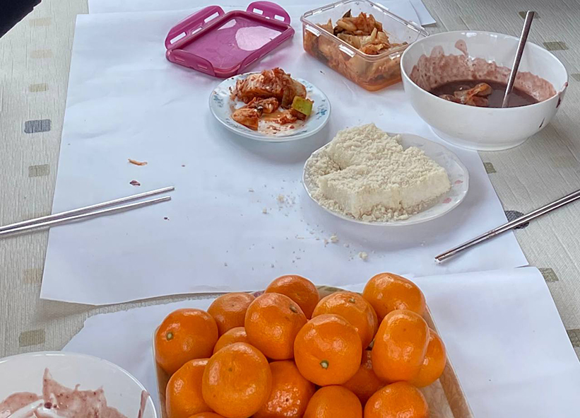 바닥 매트에 흰 종이가 넓게 깔려있고 그 위에 팥죽이 담긴 대접, 김치 접시, 흰 떡 접시, 귤이 가득 담긴 쟁반, 수저 등이 놓여있는 모습을 촬영한 사진이다. 