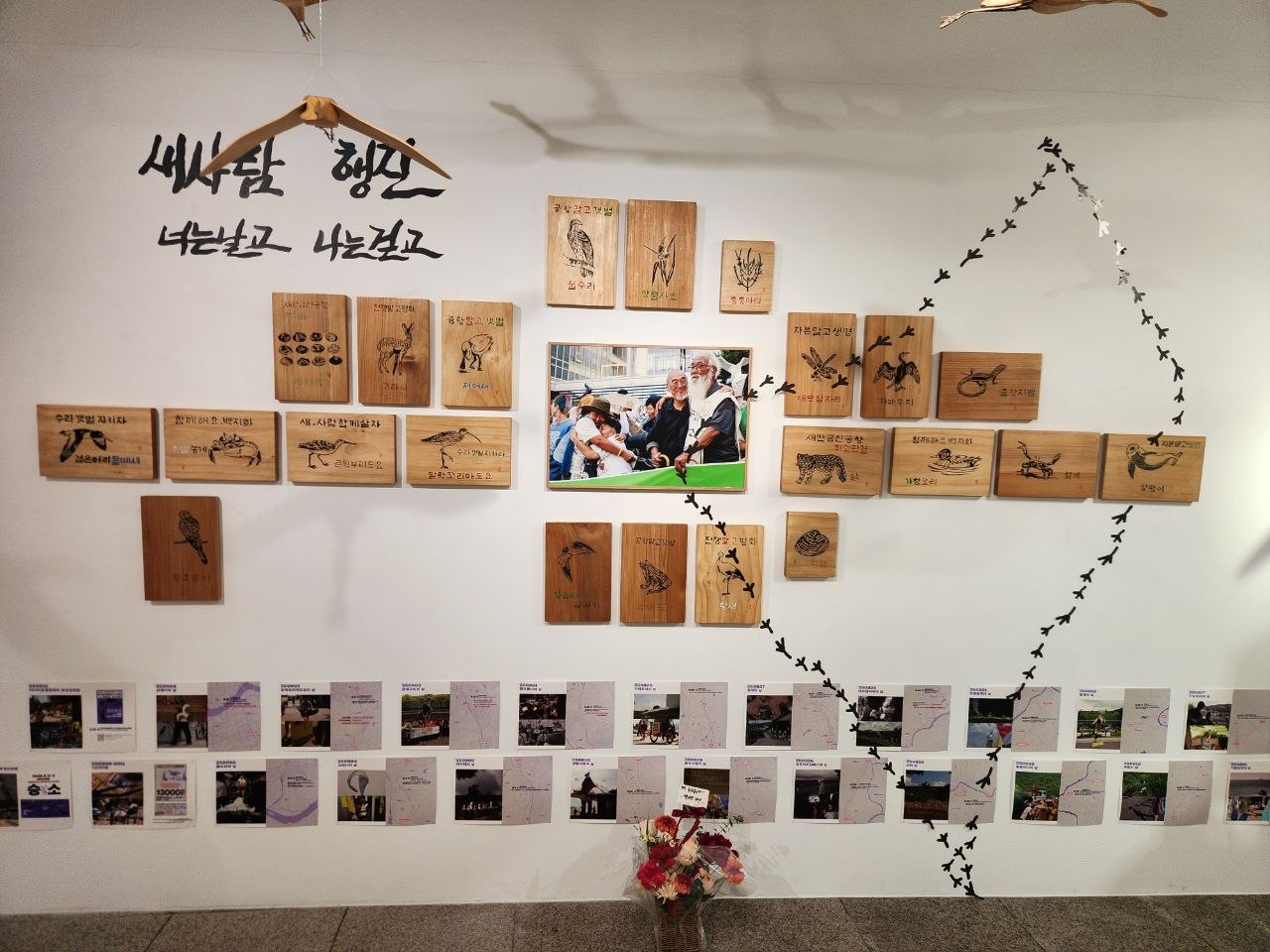 전시장 내부 벽면을 촬영한 사진이다. '새사람 행진. 너는 날고 나는 걷고'라는 문구가 왼쪽 위에 적혀있고 기록 사진과 갯벌의 생물들을 표현한 서각이 배치되어 있다. 가운데 바닥에는 꽃다발이 놓여있다. 