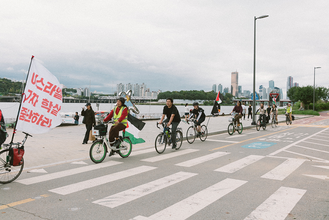 한강 공원 자전거 도로를 약 8명의 사람들이 팔레스타인 연대를 표현하는 다양한 깃발 등을 자전거에 매달고 달리는 모습을 촬영한 사진이다. 어떤 자전거에는 팔레스타인 국기가, 어떤 자전거에는 연대 머리띠가, 어떤 자전거에는 '이스라엘 가자 학살 중단하라'라는 문구가 적힌 깃발이 부착되어 있다. 자전거 도로 옆 넓은 공터에 걸어다니는 사람들이 있다. 강변에는 선착장이 있고 강 건너편에는 높은 빌딩과 숲이 보인다. 