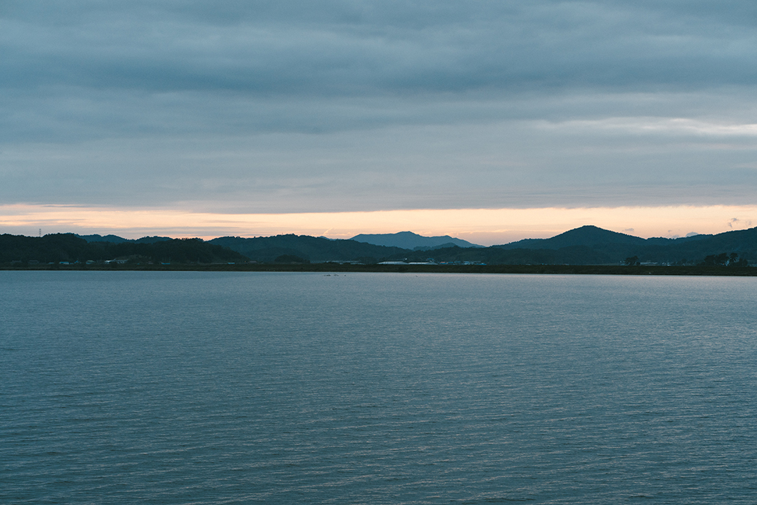 어둑어둑한 강의 모습을 촬영한 사진이다. 하늘에는 구름이 짙게 드리웠고 노을이 지는 듯하다. 멀리 산이 보인다. 