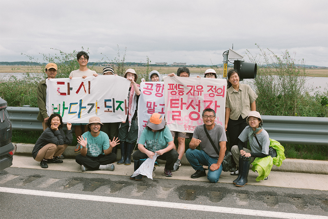 뒤로 갯벌이 보이는 도롯가에 13명의 사람들이 나란히 앉거나 서서 현수막을 들고 촬영한 사진이다. 사람들은 웃는 얼굴로 주먹을 들거나 손을 펼치고 있다. 서 있는 사람들이 든 현수막은 3개이며 왼쪽 현수막부터 각각 '다시 바다가 되자', '공항 말고 공존(글씨가 가려져서 전부 보이지 않음)', '평등, 자유, 정의, 탈성장. 핫핑크돌핀스(글씨가 가려져서 전부 보이지 않음)'이라고 적혀있다. 도롯가에는 풀숲이 우거져있고 뒤쪽 갯벌 너머로 멀리 육지의 모습이 보인다. 