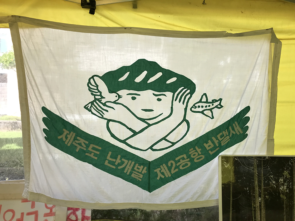 천막 내부에 현수막이 걸려있는 모습을 찍은 사진이다. 노란색 텐트천 앞쪽에 흰색 천으로 된 현수막이 있고, 현수막에는 산 모양 머리를 한 사람이 한 손으로는 새를 안고 한 손으로는 비행기를 막는 모습의 그림과 '제주도 난개발, 제2공항 반댈새'라는 문구가 담겨있다. 아래쪽으로 피켓이 세워져있다. 