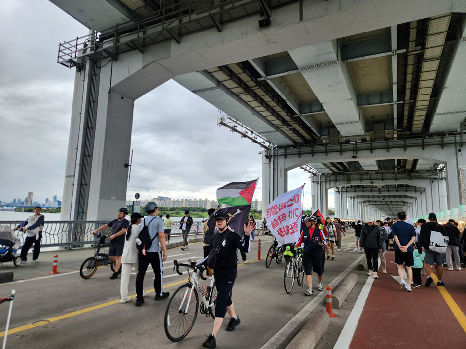 잠수교에 많은 보행자가 오가는 사이로 팔레스타인 깃발 등을 매단 자전거를 끌고 여러 명의 사람들이 걸어오는 모습을 촬영한 사진이다.
