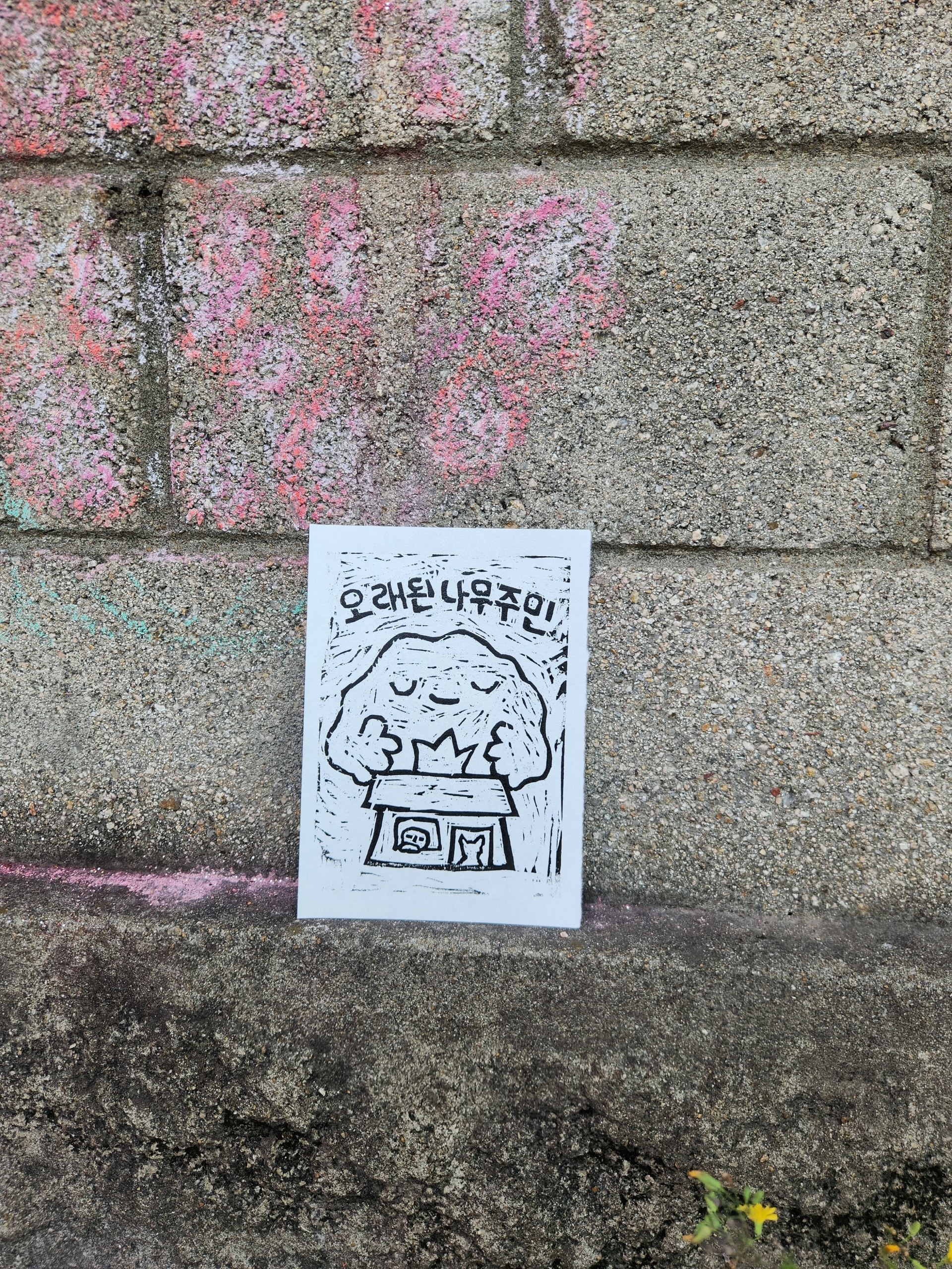 판화를 담벼락에 세워놓고 촬영한 사진이다. 집을 감싸고 있는 큰 나무의 모습, '오래된 나무주민'이라는 문구가 판화에 표현되어 있다.