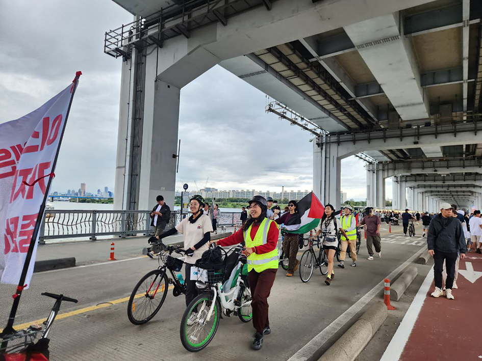 잠수교에 많은 보행자가 오가는 사이로 팔레스타인 깃발 등을 매단 자전거를 끌고 여러 명의 사람들이 걸어오는 모습을 촬영한 사진이다.