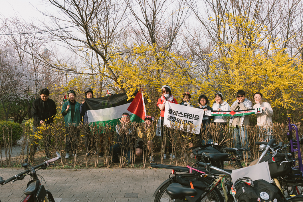 열두 명의 사람들이 개나리 나무 앞에 나란히 서있는 모습을 촬영한 사진이다. 사람들은 '팔레스타인은 해방되리라'라는 문구가 적힌 깃발과 팔레스타인 국기 등을 들고 서 있으며 앞쪽에는 자전거가 세워져있다.