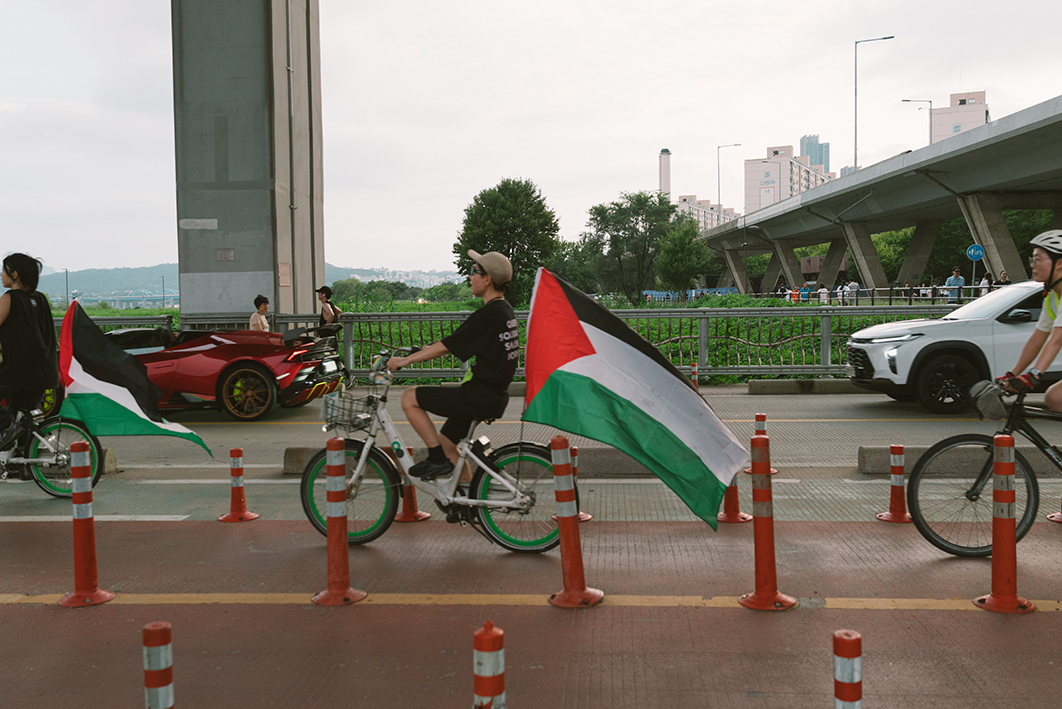 강을 가로지르는 자동차 도로 옆 자전거 도로에서 세 사람이 자전거를 타고 오른쪽에서 왼쪽으로 이동하는 모습을 촬영한 사진이다. 자전거마다 팔레스타인 깃발 또는 팔레스타인과 연대하는 메시지를 표현한 배너가 부착되어 있다. 도로에는 자동차 두 대가 자전거와 같은 방향으로 주행하고 있으며 옆 보행자 도로에는 걸어다니는 사람의 모습이 보인다. 도로 뒤편으로 한강다리에 진입하는 고가 도로, 강변의 아파트, 우거진 수풀과 나무가 무성한 공원, 멀리 강 건너편의 풍경이 보인다. 
