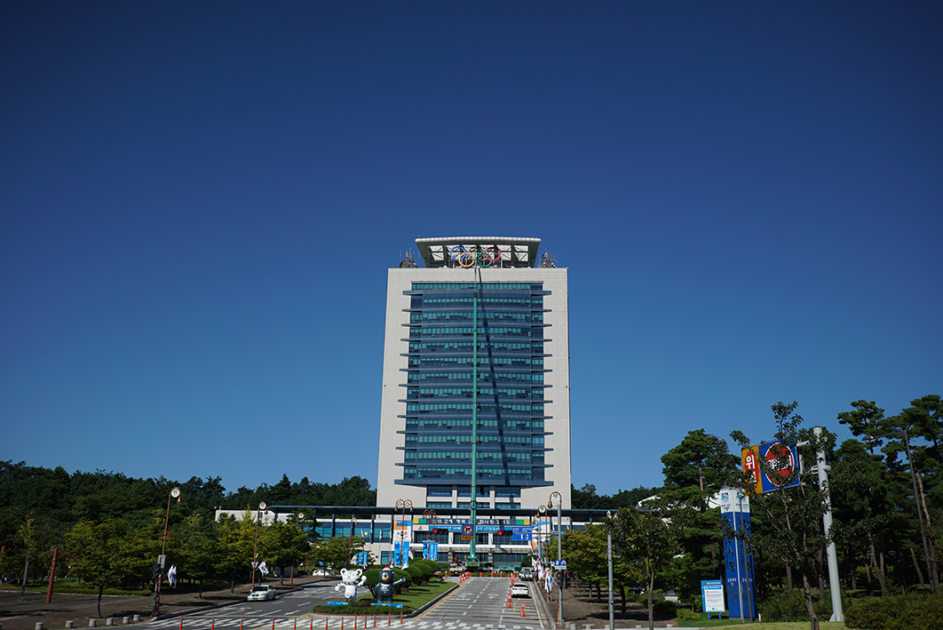 14층 높이 건물의 꼭대기에 오륜기 형상의 조형물을 설치하고 있는 장면의 사진이다. 건물 진입로 앞에는 올림픽 마스코트 조형물이 설치되어 있고 좌우로는 나무가 우거져있다. 