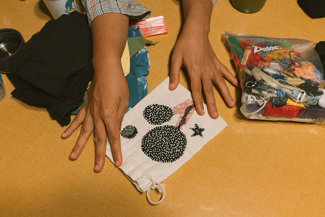 탁자 위에 여러 실과 바느질 도구가 담긴 비닐봉지, 조각 천, 수선한 작은 주머니 등이 놓여있고 한 사람이 작은 주머니를 두 손으로 펼쳐 만지는 모습을 촬영한 사진이다. 