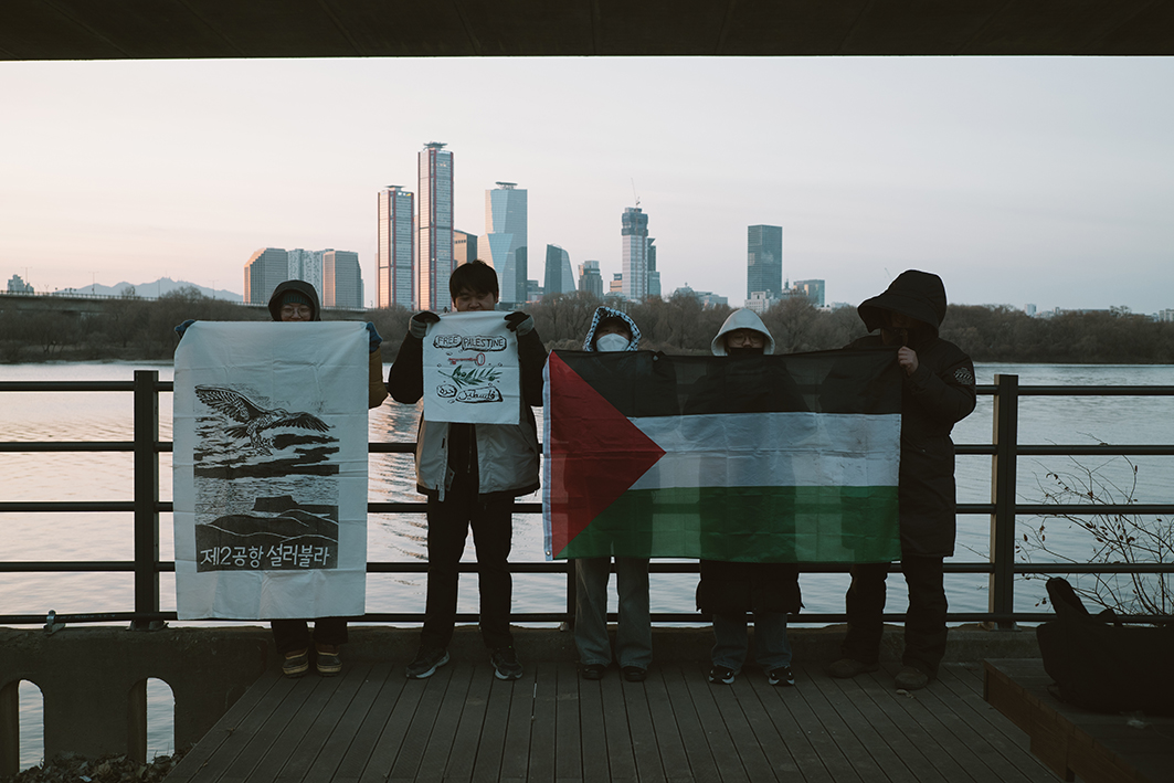 강변 난간 앞에서 다섯 사람이 손에 자란히 서서 배너나 팔레스타인 국기를 들고 서 있는 모습을 촬영한 사진이다. 팔레스타인 해방과 제주 제2공항 반대 메시지가 배너에 표현되어 있다. 뒤로는 밤섬과 여의도 빌딩의 모습이 보인다.