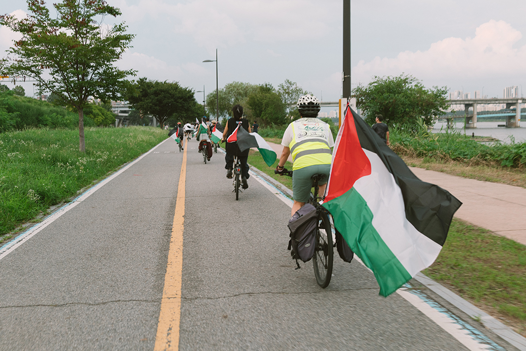 한 무리의 사람들이 한강 자전거 도로를 달리는 모습을 촬영한 사진이다. 자전거마다 팔레스타인 깃발 또는 팔레스타인과 연대하는 메시지를 표현한 배너가 부착되어 있다. 자전거 도로 오른편 보행자 도로에는 보행자들이 있고 좌우로는 우거진 수풀과 나무들이 있다. 오른편으로 한강과 한강대교, 강 건너의 풍경이 보인다. 하늘은 맑고 다소 구름이 껴 있다. 