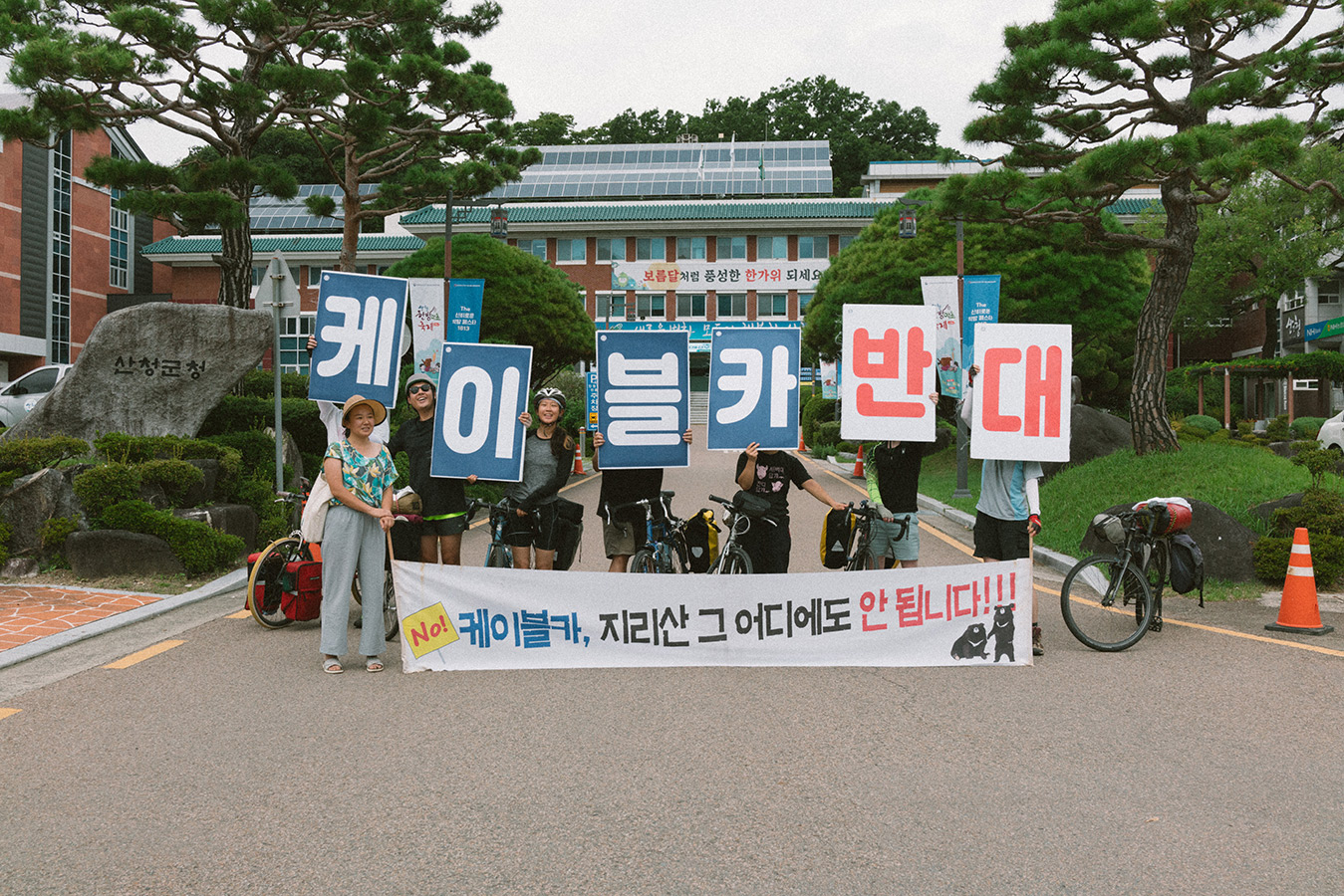 관공서 건물 앞 진입 도로에서 여러 사람들이 피켓과 배너를 들고 있다. 피켓에는 '케이블카 반대', 배너에는 '케이블카, 지리산 그 어디에도 안됩니다!!!' 라고 적혀있고 사람들 옆에는 자전거 몇 대가 있다.