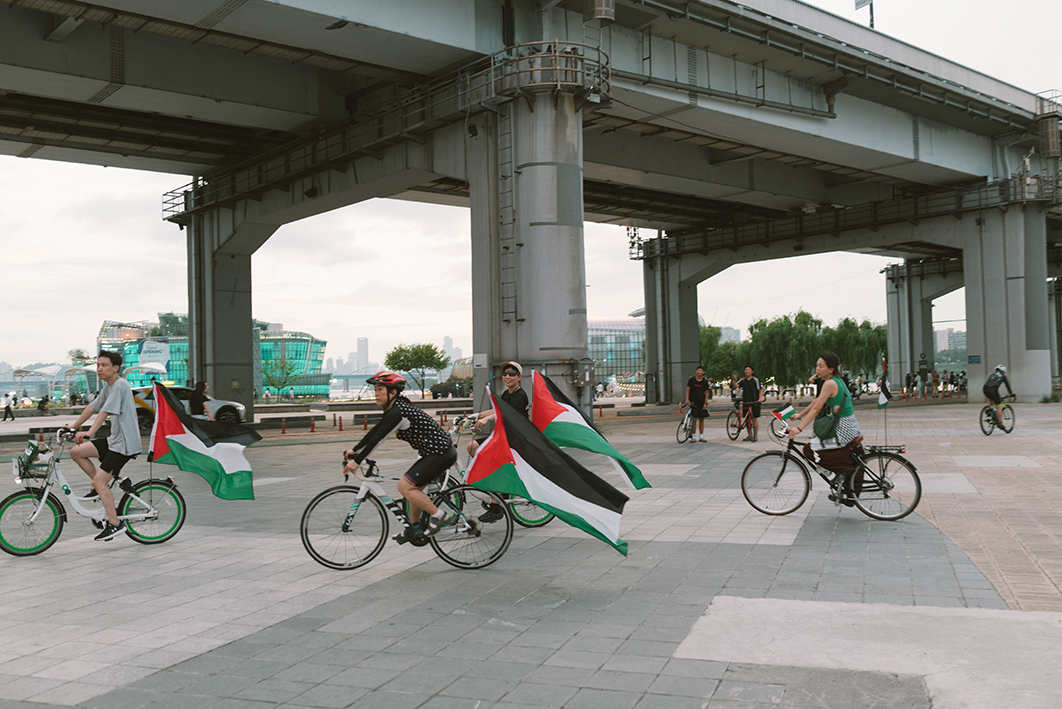 네 명의 사람들이 한강 공원 공터에서 자전거를 타고 있는 모습을 촬영한 사진이다. 자전거마다 팔레스타인 깃발 또는 팔레스타인과 연대하는 메시지를 표현한 배너가 부착되어 있다. 뒤로는 강을 가로지르는 다리, 한강변의 여러 시설물, 자전거를 타거나 산책하는 여러 사람들의 모습이 보인다.