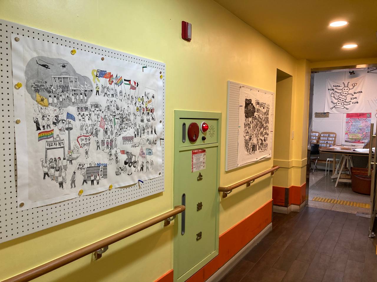 복도 한쪽 벽면 타공판에 같은 장면을 담은 드로잉과 판화가 붙어있는 모습을 촬영한 사진이다. 두 타공판 사이에는 소화전에 위치해있고 타공판 아래에는 손잡이가 있다. 오른쪽 타공판 오른편에는 다른 출입구가 있고 복도 끝쪽 문은 안쪽으로 열려있다. 복도 너머의 공간에는 탁자와 진열대 등이 보인다. 