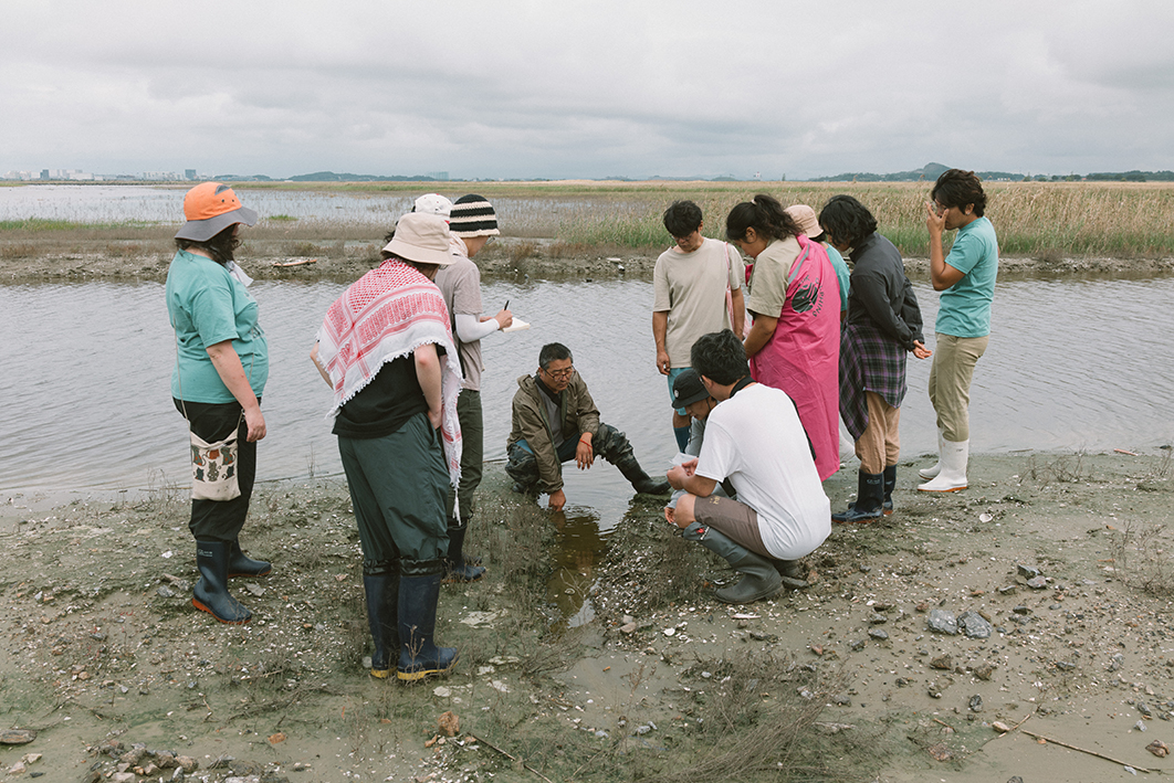  갯벌 물길 옆에서 약 11명의 사람들이 둥글게 둘러 서거나 쪼그려 앉아 있는 모습을 촬영한 사진이다. 가운데에 한 사람은 물길 위에 걸터앉아 한쪽손을 물에 담그고 있으며 나머지 사람들은 그를 쳐다보고 있다. 물길 뒤편으로는 풀이 자란 너른 갯벌의 모습, 멀리 육지의 모습이 보인다. 