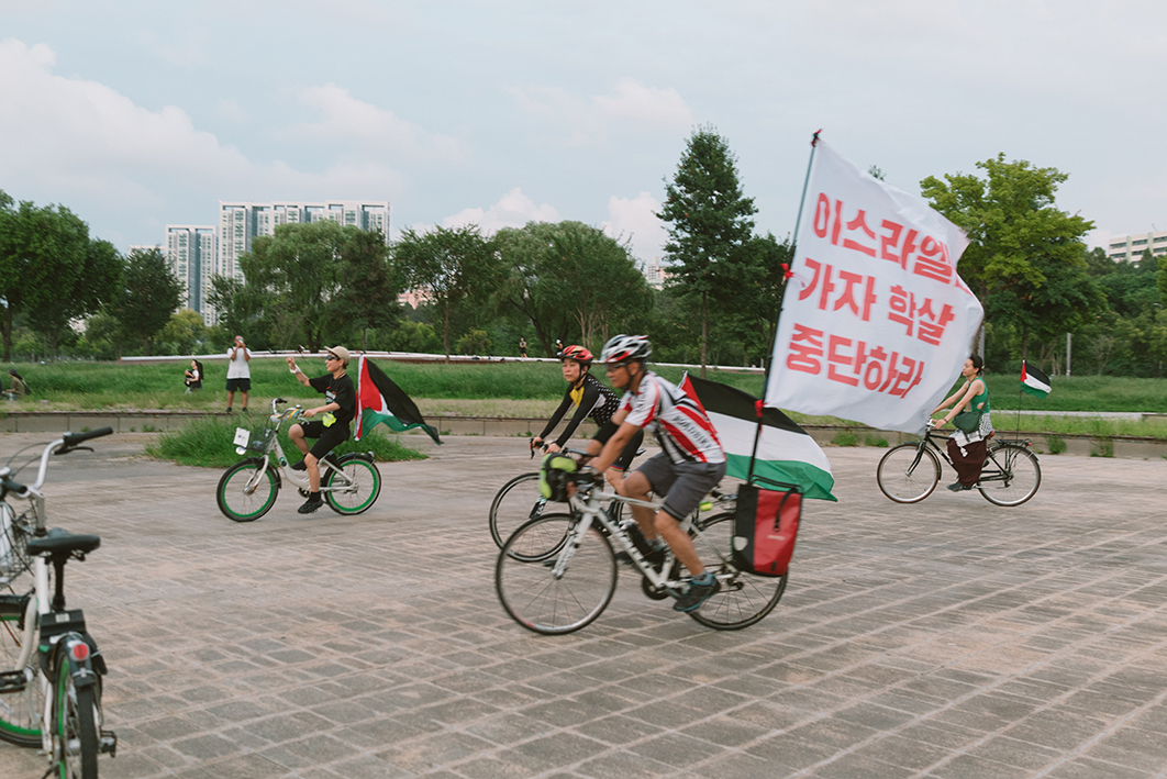 네 명의 사람들이 한강 공원 공터에서 자전거를 타고 있는 모습을 촬영한 사진이다. 자전거마다 팔레스타인 깃발 또는 팔레스타인과 연대하는 메시지를 표현한 배너가 부착되어 있다. 가장 가까운 곳에서 주행 중인 자전거에 부착된 흰색 깃발에는 붉은색 글씨로 '이스라엘은 가자 학살 중단하라'라고 적혀있다. 수풀과 나무가 우거진 뒤쪽 공원 보행로로 지나가는 사람들이 보이고, 멀리 아파트가 있다. 하늘은 맑고 다소 구름이 껴 있다.
