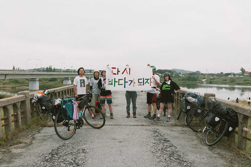 강을 가로지르는 작은 다리 위에 여섯 명의 사람들이 서서 '다시 바다가 되자'라는 현수막을 들고 정면을 바라보고 서 있다. 그 옆에는 자전거들이 세워져있다. 뒤로는 강가와 또 다른 다리, 마을의 모습이 보인다. 