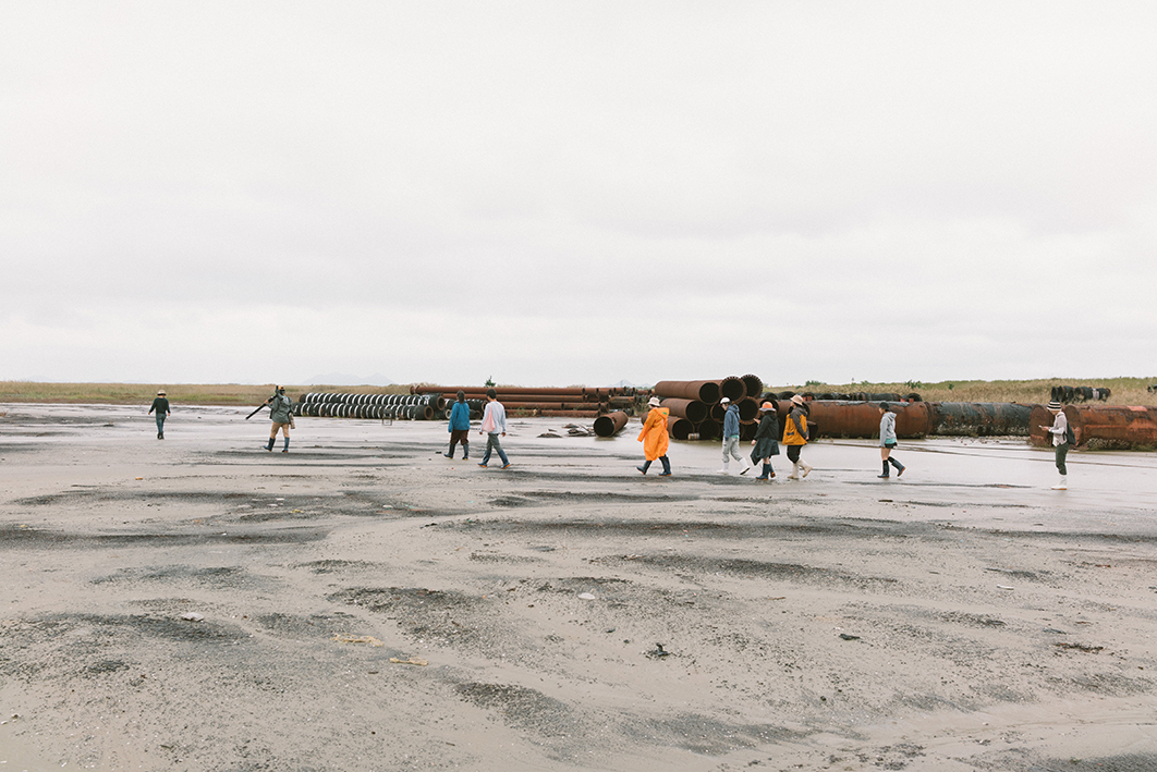 갯벌에서 10명의 사람들이 띄엄띄엄 오른쪽에서 왼쪽으로 걸어가는 모습을 촬영한 사진이다. 사람들이 걸어가는 옆으로 거대한 파이프 등의 자재가 쌓여있다. 갯벌 먼 쪽에는 갈대 등 식물이 우거져있다. 