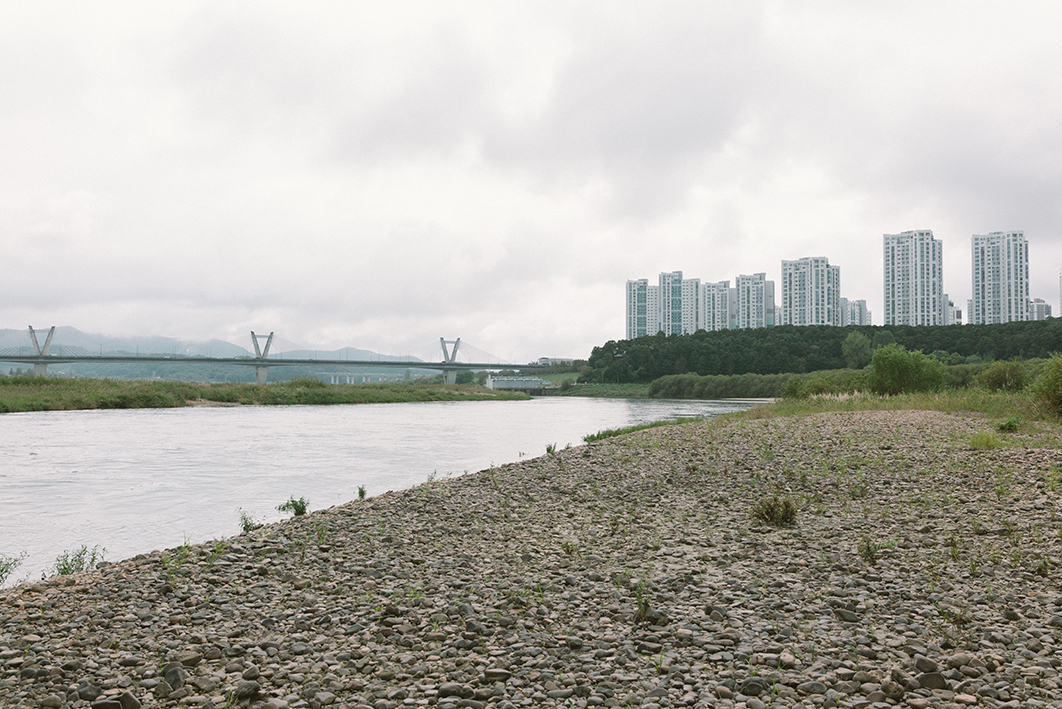 강과 강가, 그 옆의 아파트 단지, 그리고 강을 가로지르는 보의 모습을 촬영한 사진이다. 강가에는 자갈과 수풀이 넓게 자리잡고 있고, 강은 오른쪽 열린 수문 방향으로 흘러간다. 