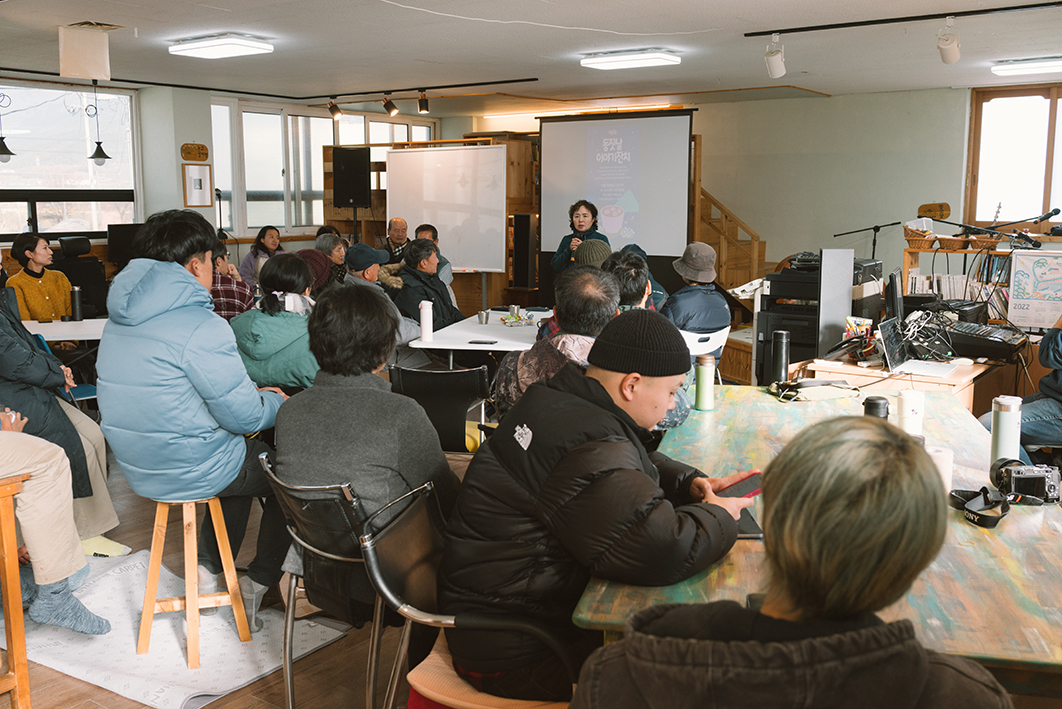 탁자와 의자 등이 있는 실내 공간에 스무 명 남짓의 사람들이 앉아 있고 앞쪽 중앙에는 프로젝터 화면이 나오는 스크린과 마이크를 든 한 사람이 있으며, 오른쪽 편에는 악기와 마이크 등의 공연 장비가 놓여있는 모습을 촬영한 사진이다. 마이크를 든 사람은 중년의 여성이며 의자에 앉은 사람들 대부분은 그 사람의 이야기에 귀를 기울이는 듯한 모습이다. 다수의 사람들은 두꺼운 겨울 옷을 입고 있다. 