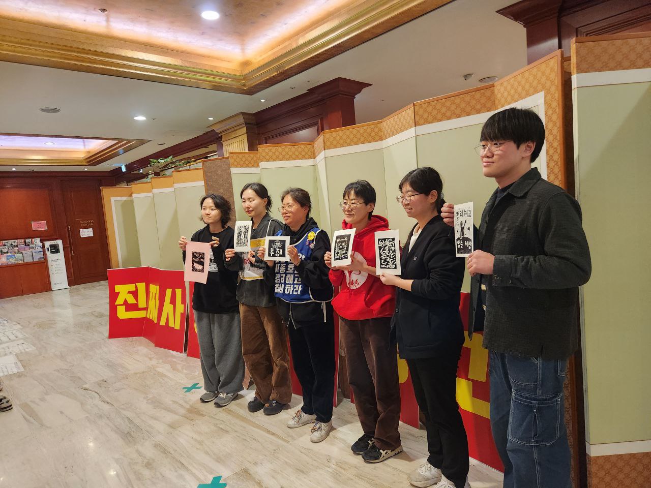 6명의 사람들이 실내의 병풍과 피켓 앞에 나란히 서서 판화를 들고 있는 모습을 촬영한 사진이다. 
