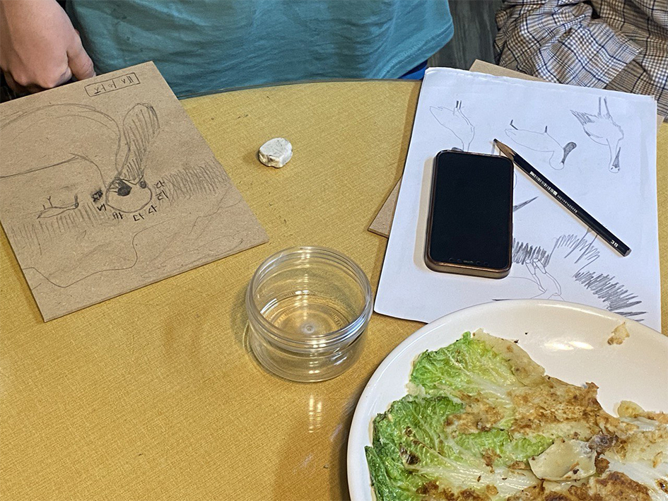 탁자 위에서 각각 목판과 종이에 스케치하는 모습을 촬영한 사진이다. 목판에는 저어새의 모습이, 종이에는 황새와 저어새의 모습이 그려져있다. 주변에는 지우개와 연필, 핸드폰, 접시에 담긴 배추전 등이 놓여있고 탁자 옆에 앉은 사람들의 모습이 일부 보인다. 