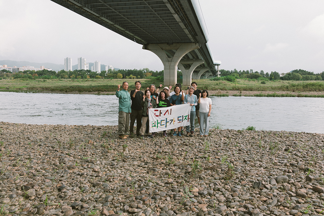 자갈이 많은 너른 강가에 약 11명의 사람들이 '다시 바다가 되자'라는 현수막을 들고 서서 한쪽 주먹을 들고 외치는 모습을 촬영한 사진이다. 뒤로는 강과 강변에 우거진 수풀, 그 강을 가로지르는 다리, 멀리 도시의 모습이 보인다. 