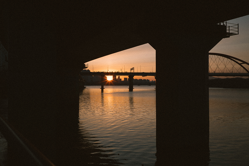 다리 기둥 사이로 강가로 떠오르는 해를 촬영한 사진이다. 강건너편에 있는 크고 작은 빌딩, 강 중앙을 가로지르는 다리의 실루엣이 선명하게 보이고, 강물에는 햇빛이 비춰지기 시작한다.