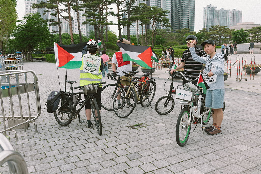 길 위에 자전거 다섯대와 네 사람이 서서 주먹을 들거나 자전거에 부착된 팔레스타인 국기를 펼쳐들고 있는 장면의 사진이다. 일부 사람들의 옷에는 팔레스타인 연대의 메시지를 담은 배너가 달려있고, 오른편에 모자를 쓴 사람은 쿠피예를 두르고 있다.