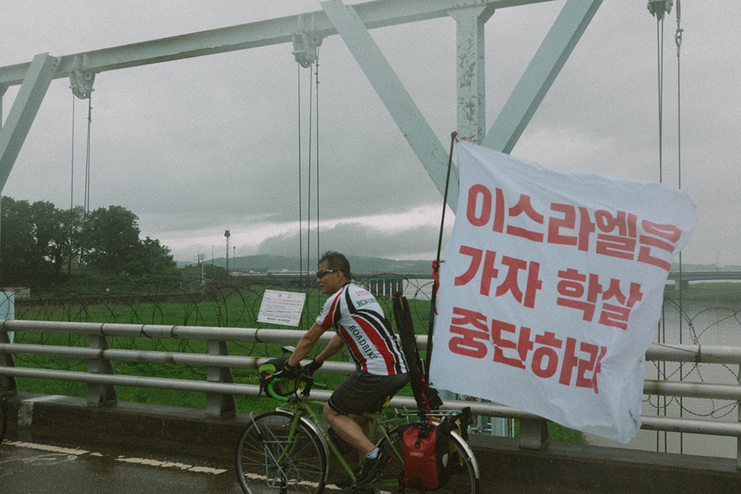 다리를 건너는 자전거의 모습을 촬영한 사진이다. 자전거에는 '이스라엘은 가자 학살 중단하라'라고 적힌 큰 깃발이 부착되어 있다. 다리에는 철조망과 군사구역임을 알리는 경고판이 붙어있다. 멀리 강 옆으로 습지와 흐린 하늘이 보인다.